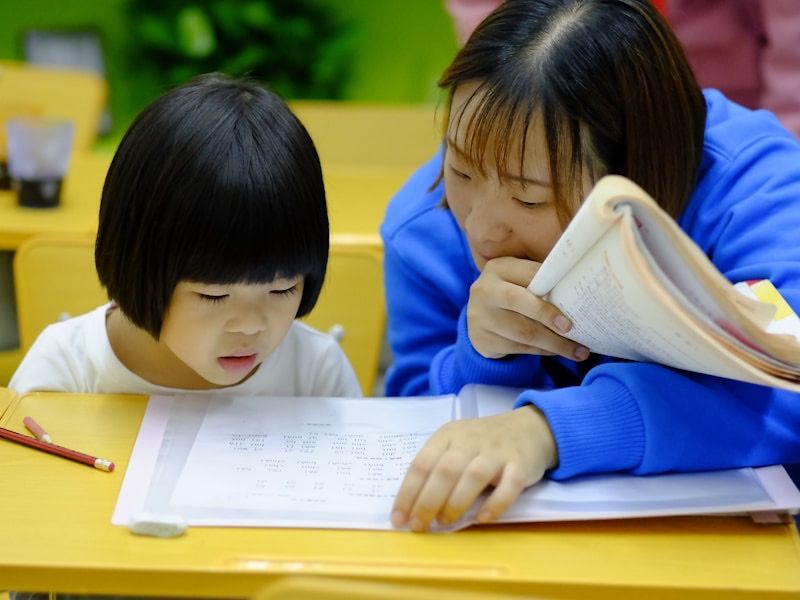 A teacher and a young student reading a book together at a yellow desk